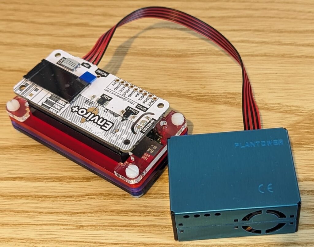 Image shows a Raspberry Pi environmental monitoring unit which includes a Raspberry Pi in a case, the Enviro+ sensors on top, connected via a cable is the particulate matter sensor.